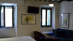 A hotel room with a bed, a desk, a chair, a sofa, a TV, and two windows with blinds. - Casa Messi - Casa Messi a Vicolo Della Rivolta 31 Apt 2 (Pitigliano)