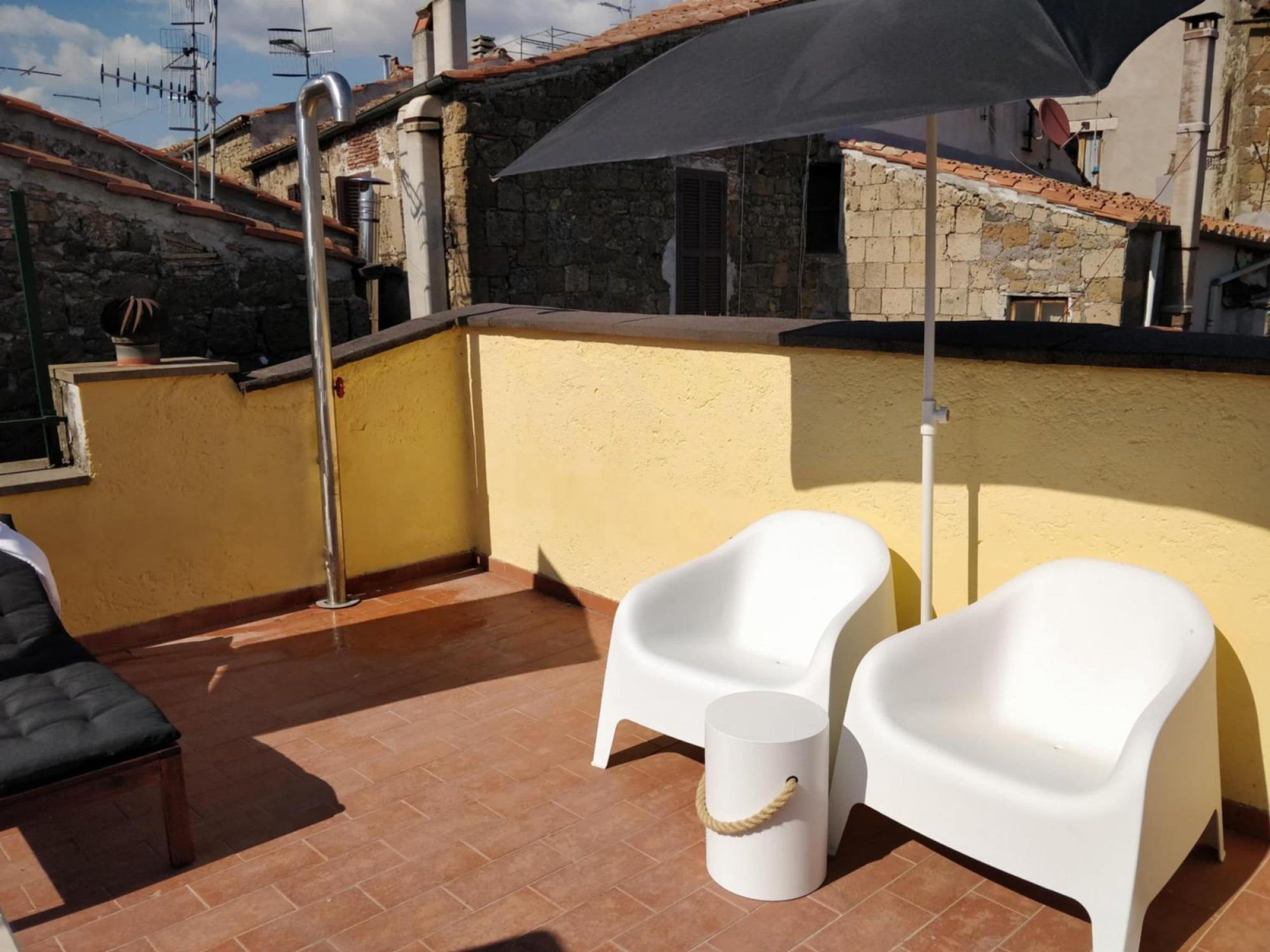 Une terrasse sur le toit avec deux chaises en plastique blanches, un petit tabouret blanc et un grand parasol noir.