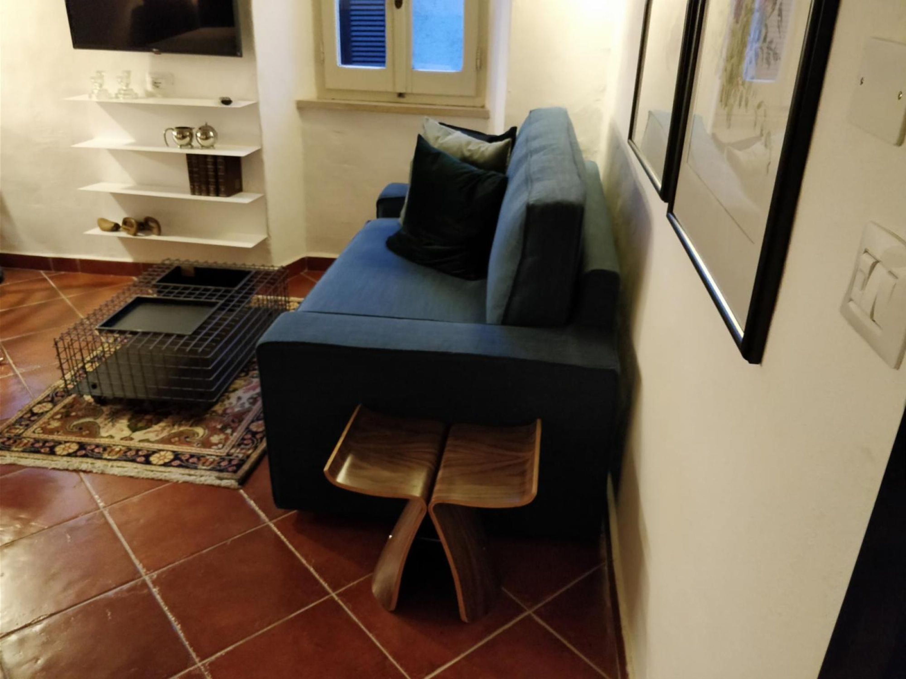 A living room with a dark blue sofa, a wooden bench, a TV, a window, and a tiled floor.