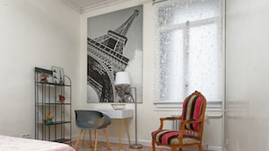 Ein Zimmer mit einem Bett, einem Schreibtisch mit Stuhl, einem großen Fenster mit Spitzenvorhängen und einem an der Wand befestigten Regal mit Dekorationen.
