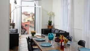 Ein Essbereich mit einem langen Holztisch, gedeckt für sechs Personen, weißen Stühlen, einer schwarzen Couch und Blick auf die Stadt durch große Fenster.