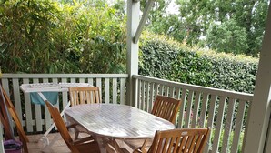 Une terrasse couverte avec une table et des chaises en bois, entourée d’une clôture blanche et de verdure.