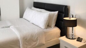 A neatly made bed with white bedding and a black headboard, a bedside table with a lamp, and a small drawer unit.