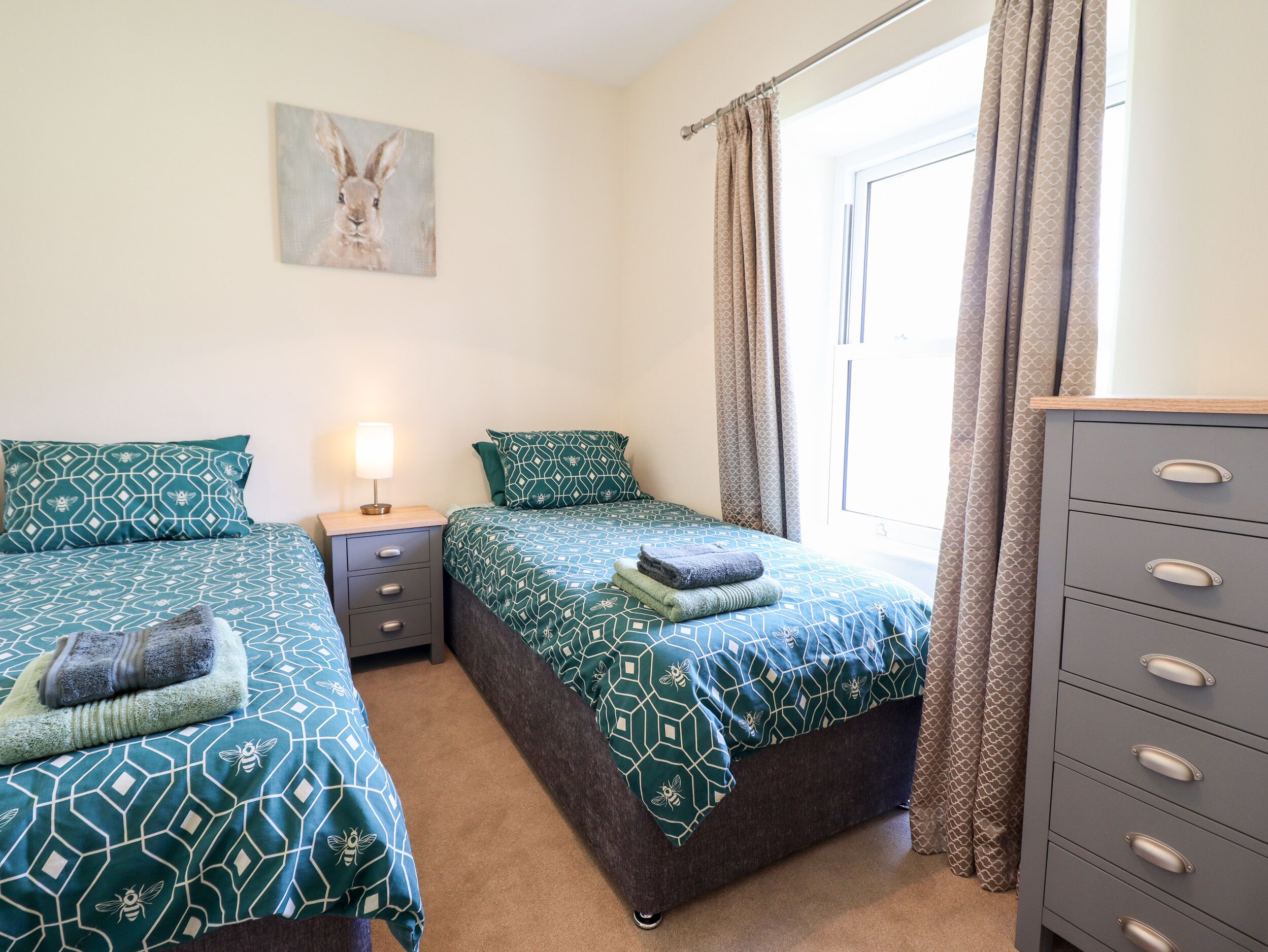 Une chambre avec deux lits, une table de chevet avec une lampe, une commode et une fenêtre avec des rideaux.
