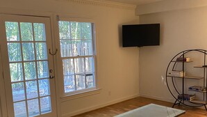 A room with a wooden floor, a glass table, a grey sofa, a TV mounted on the wall, and a bookshelf.