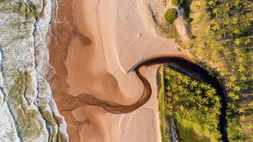 Vista aérea de uma praia com uma grande formação arenosa curva, um rio ou riacho, e vegetação densa à direita.