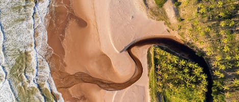 Vista aérea de una playa con una gran formación arenosa curva, un río o arroyo, y vegetación densa a la derecha.