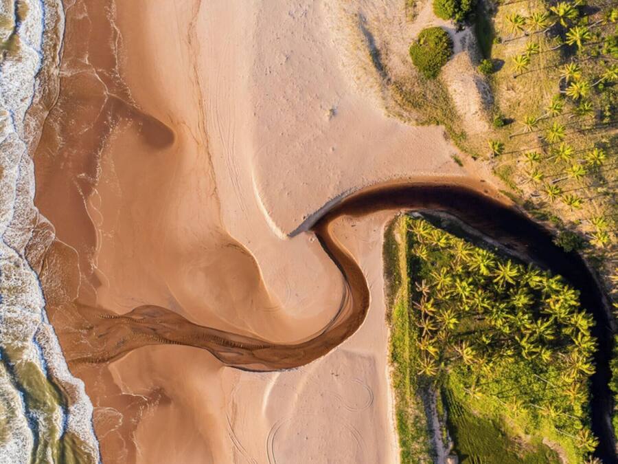 An aerial view of a beach with a large curved sand formation, a river or stream, and dense vegetation on the right.