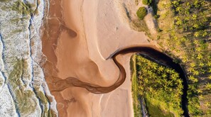 Vista aérea de una playa con una gran formación arenosa curva, un río o arroyo, y vegetación densa a la derecha.