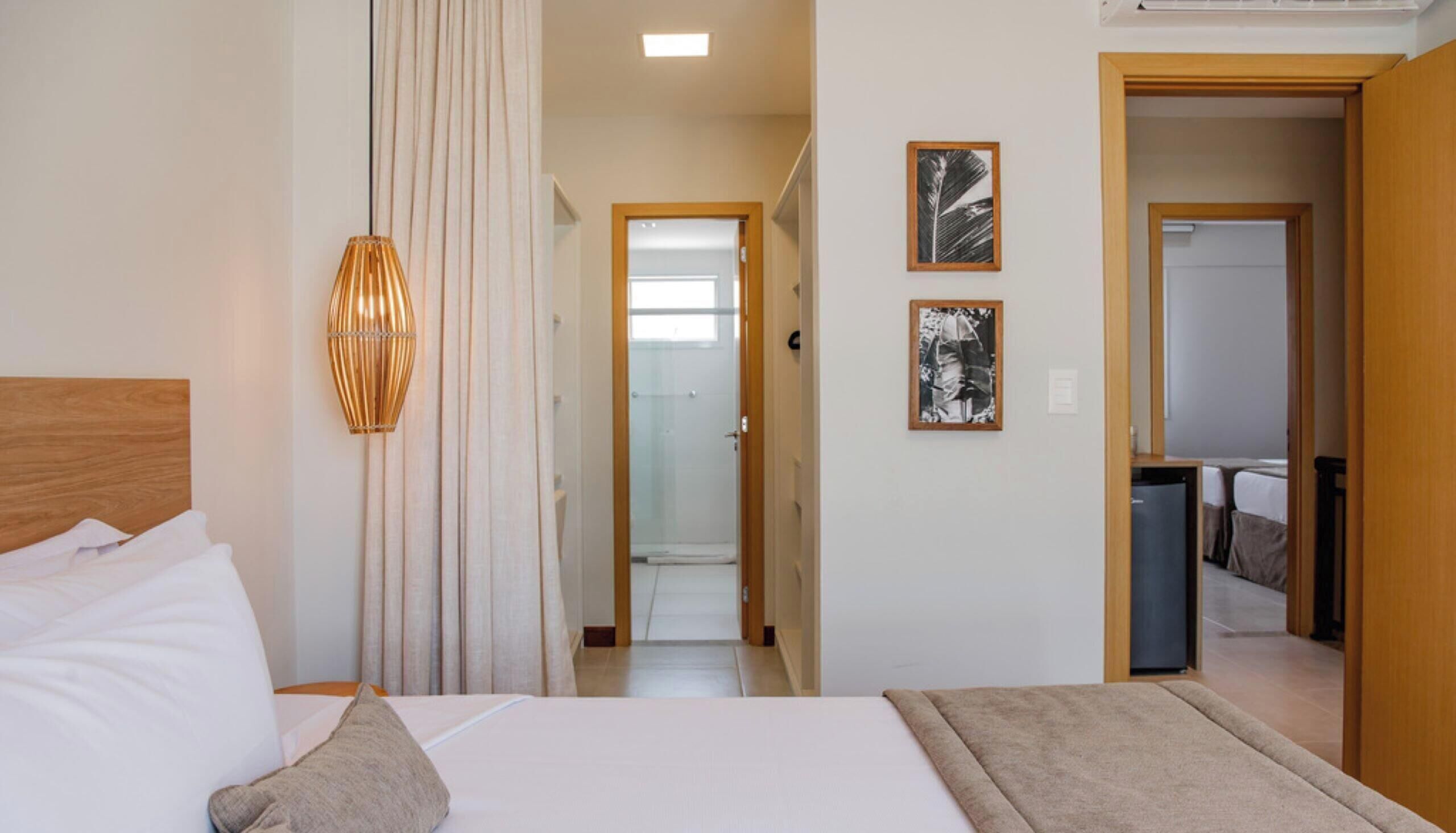 A hotel room with a bed, a bedside lamp, a door leading to a bathroom, and a hallway with a shower.