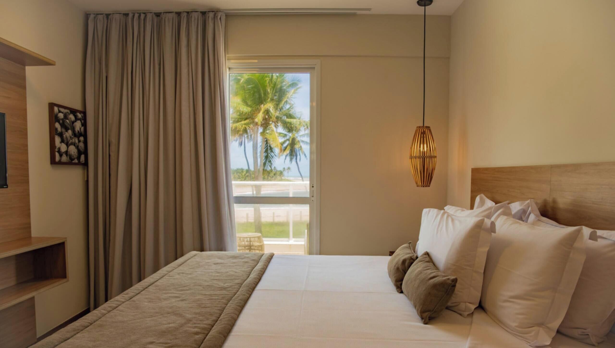 Quarto de hotel com uma cama, mesa de cabeceira, abajur e vista para palmeiras pela janela.