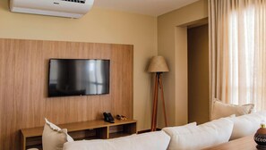 A modern hotel room with a flat-screen TV mounted on a wooden panel wall, a wooden side table with a lamp, and a sofa with a wooden coffee table. - Ponta de Inhambupe Residences (Esplanada)