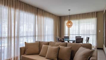 Uma sala de estar com um sofá bege, uma mesa de jantar com cadeiras e cortinas leves.