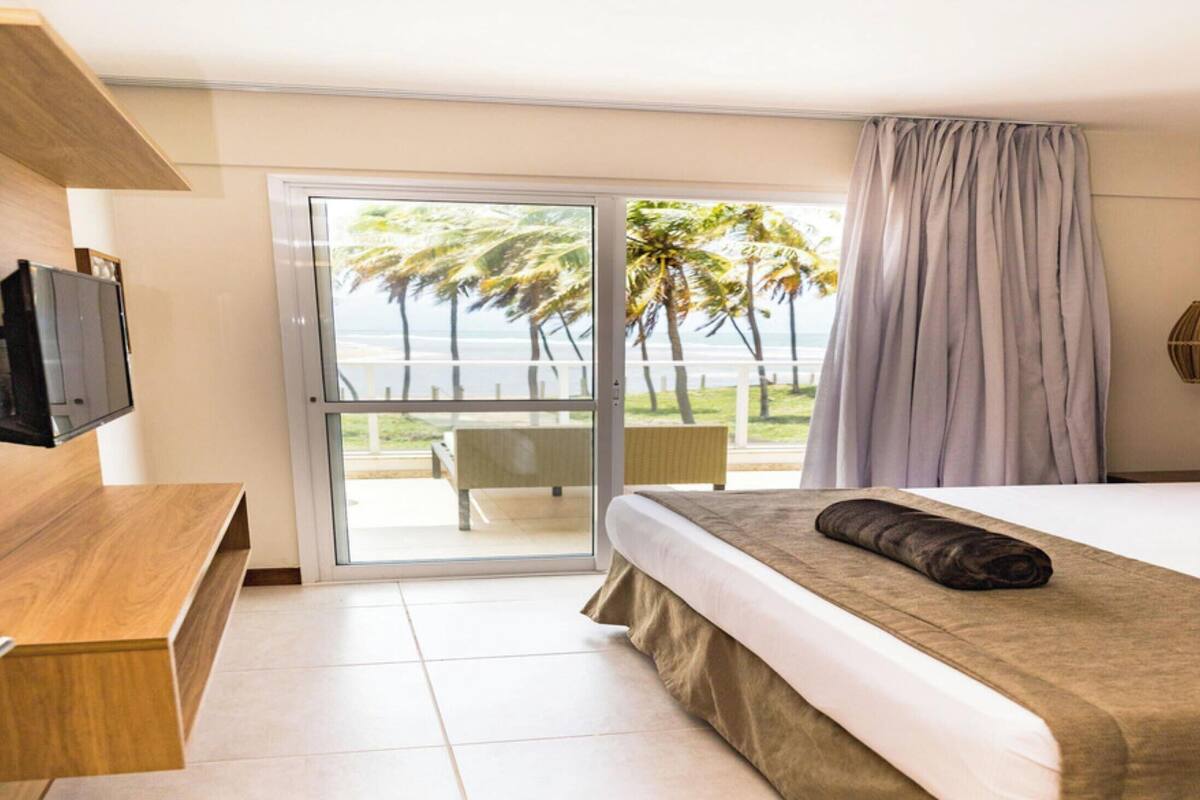 A hotel room with a bed, a TV, a balcony with a view of palm trees and the ocean.