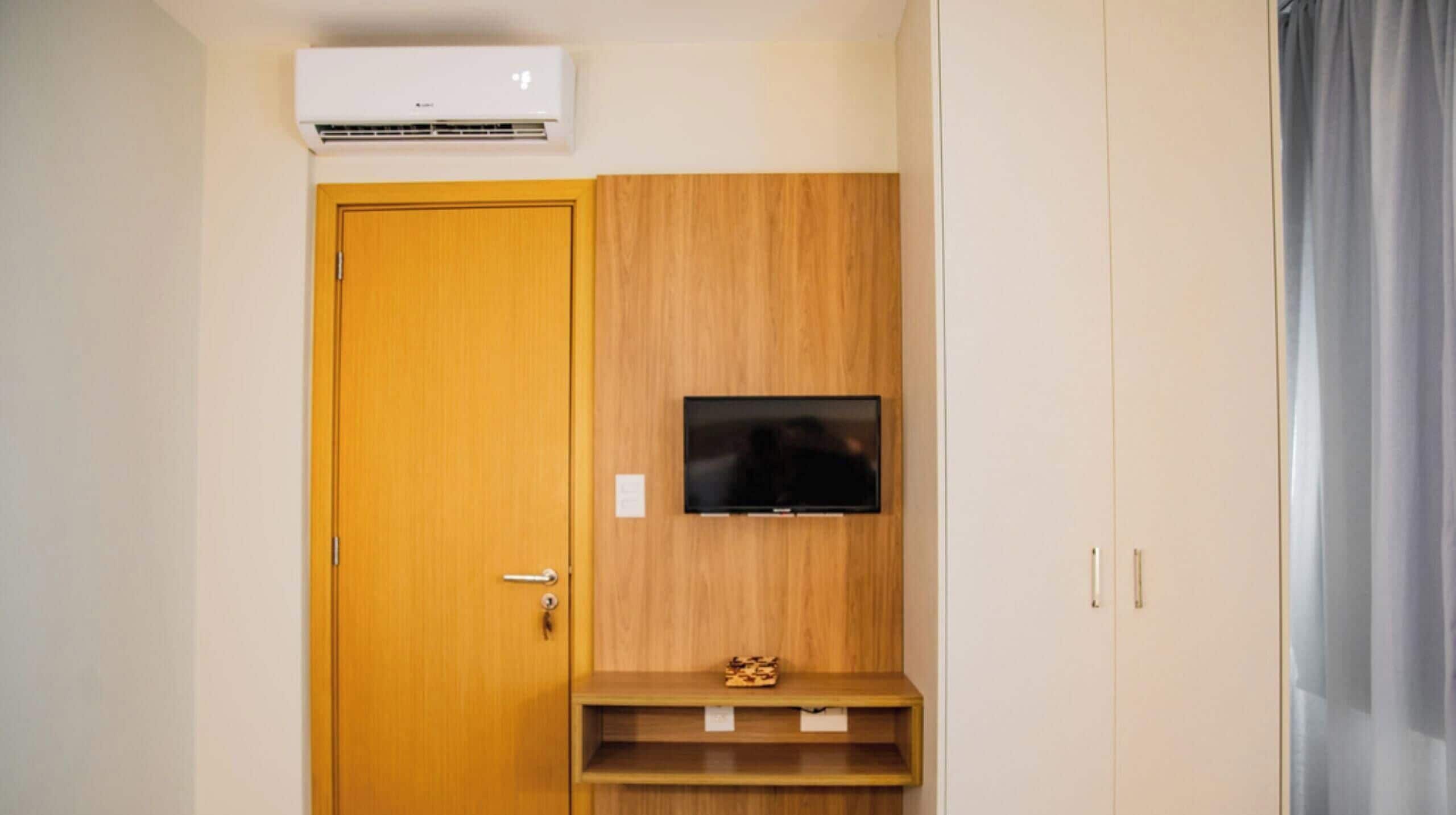 Um quarto com porta de madeira, televisão montada e uma pequena prateleira de madeira.