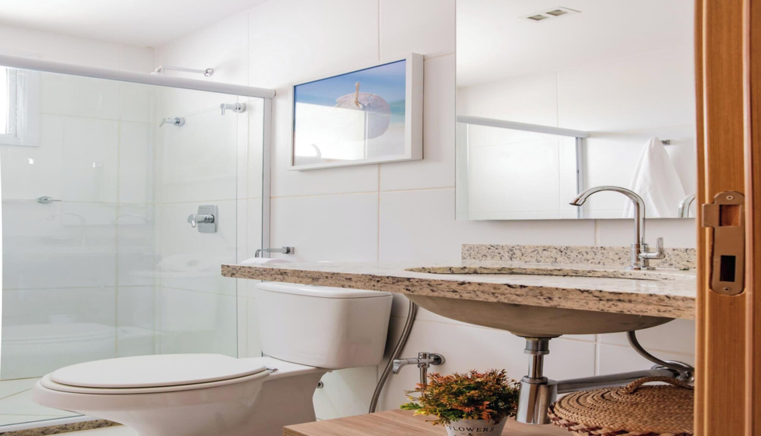 Uma casa de banho moderna com chuveiro em cabine de vidro, sanita branca, lavatório com bancada de granito e espelho sobre o lavatório.