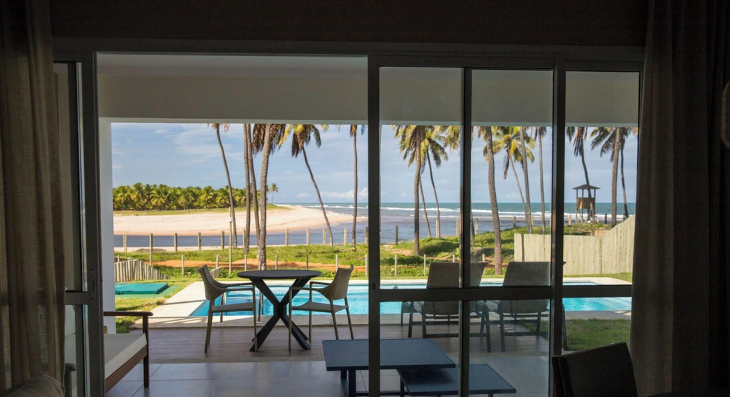Interior moderno com uma grande porta de vidro deslizante que dá para uma área de piscina com vista para a praia e palmeiras.
