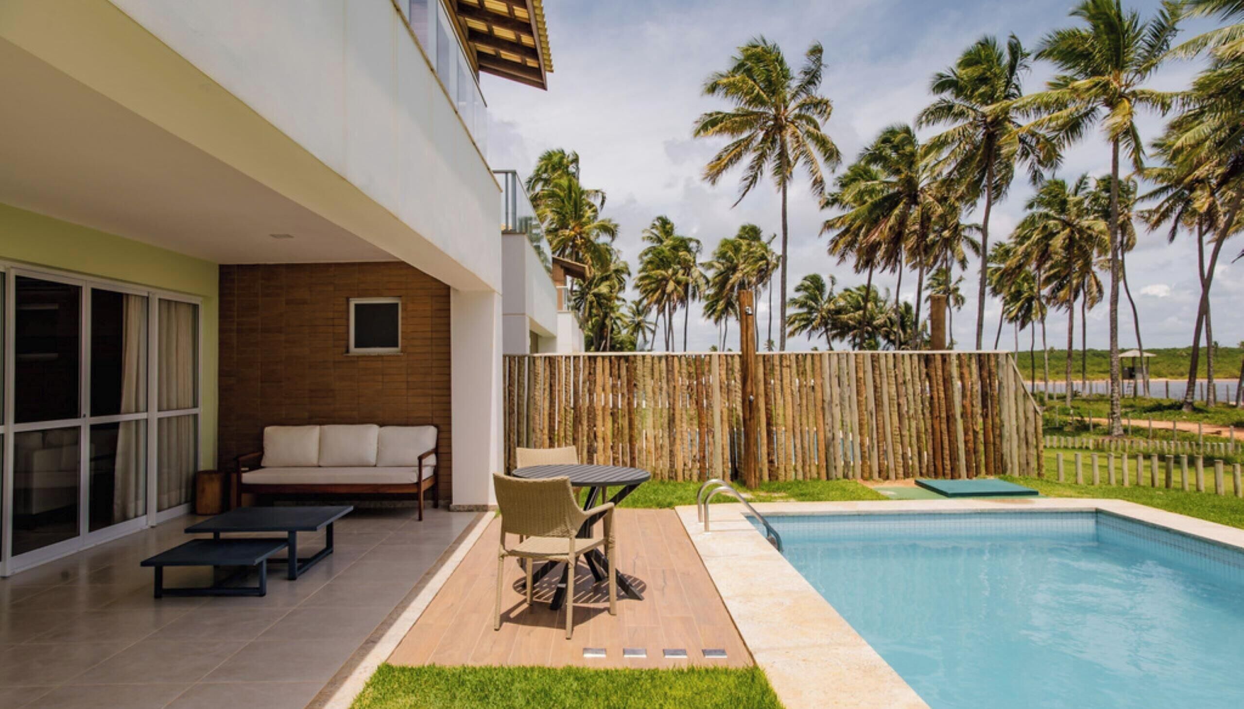 Un espacio exterior moderno con piscina, cerca de madera y palmeras al fondo.