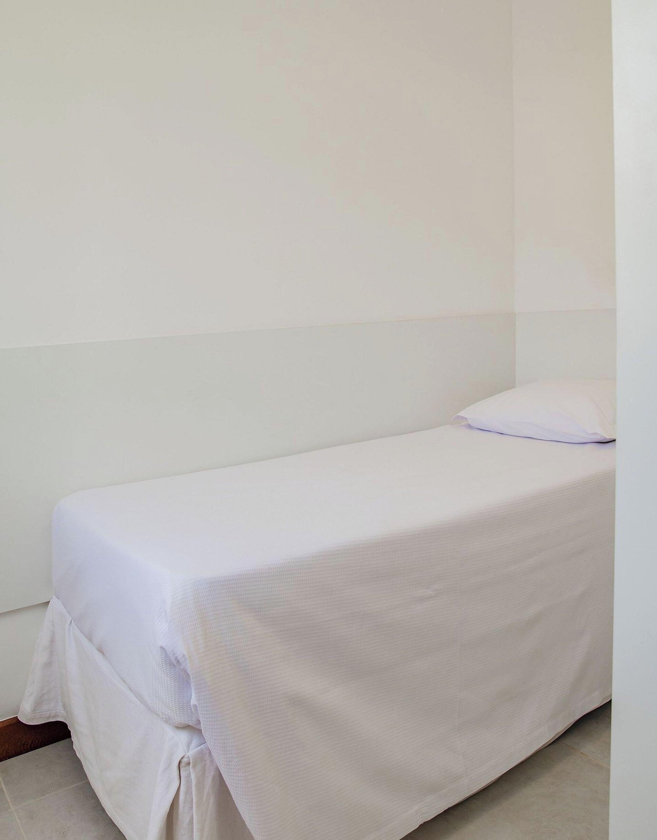 Una habitación pequeña con cama individual, una colcha blanca sencilla y una almohada.