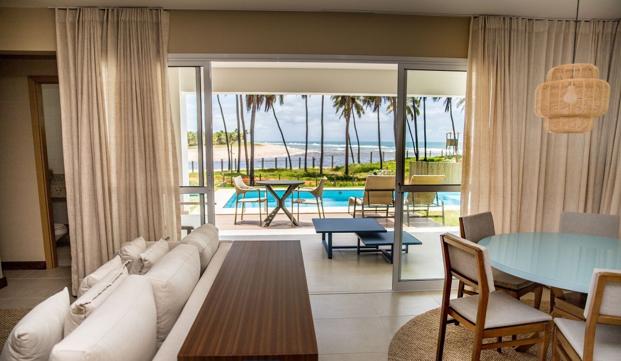 A hotel room with a sofa, a dining table, and chairs. There is a view of a pool and palm trees outside.