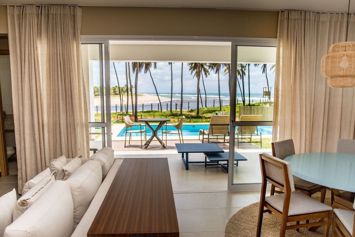 A hotel room with a sofa, a dining table, and chairs. There is a view of a pool and palm trees outside.