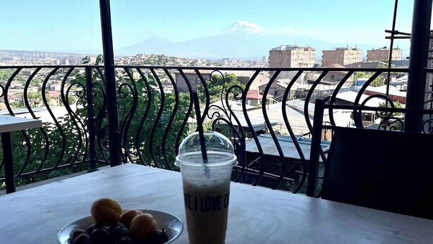 A table with a laptop, a drink, and a bowl of fruit on a balcony overlooking a cityscape and a mountain.