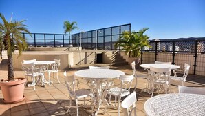 En udendørs terrasse med hvide metalborde og stole, plantekasser og havudsigt.
