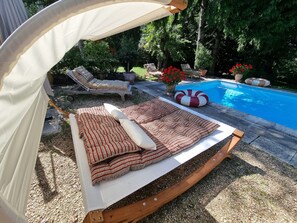 A poolside area with a wooden swing chair, a cushioned lounge chair, and a canopy. - Les Jardins Saint Eloi (Le Lonzac)