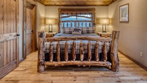 A rustic bedroom with a wooden bed frame, plaid bedding, and a large window with a view of trees.