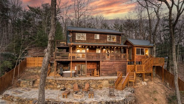 A two-story log cabin with a stone patio, outdoor seating, and a hot tub.