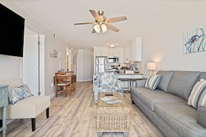 A modern living room with a sofa, a coffee table, a dining area with chairs, a kitchen with appliances, and a ceiling fan.