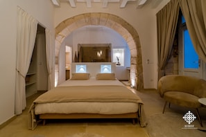 A bedroom with a large bed, an arched stone doorway, a small table, and a chair.