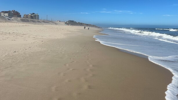 A wide sandy beach with gentle waves, a clear sky, and a few buildings in the background.