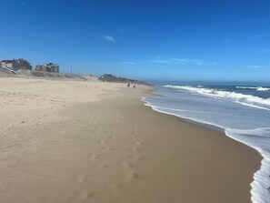 Una amplia playa arenosa con olas suaves, cielo despejado y algunos edificios al fondo.