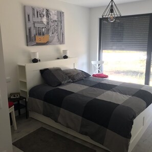 A modern bedroom with a large bed, a side table, a radio, a hanging light fixture, and a window with blinds. - La casa de férias contemporary house (Ilha)