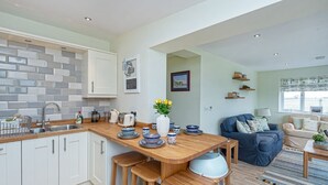 A modern kitchen with white cabinets, a wooden countertop, and bar stools. There is a dining area with a blue sofa and a coffee table. - Cryd Y Nant -  a bungalow that sleeps 6 guests  in 3 bedrooms (Rhosneigr)