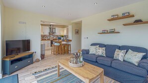 A living room with a TV, sofa, coffee table, and kitchen in the background. - Cryd Y Nant -  a bungalow that sleeps 6 guests  in 3 bedrooms (Rhosneigr)