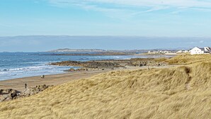 A coastal landscape with a grassy hill, rocky shoreline, and a white building with a sloped roof. - Cryd Y Nant -  a bungalow that sleeps 6 guests  in 3 bedrooms (Rhosneigr)