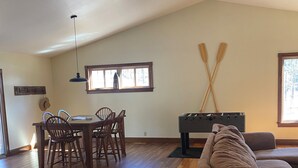 Amplia sala de estar con piso de madera, una mesa de comedor con sillas, un sofá, mesa de billar y dos palos apoyados en la pared.