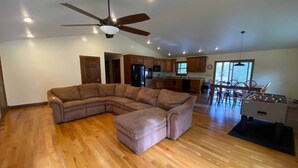 Un salón espacioso con un amplio sofá en forma de L, un ventilador de techo, suelo de madera y una cocina al fondo.