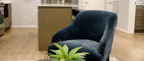 Un salon moderne avec un fauteuil bleu foncé, une table basse ronde agrémentée d’une plante, un coin cuisine avec un pare-feu en marbre et une guitare accrochée au mur.