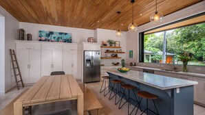 Una cocina moderna con techo de madera, armarios blancos, una isla central con taburetes y un amplio ventanal con vistas a zonas verdes.
