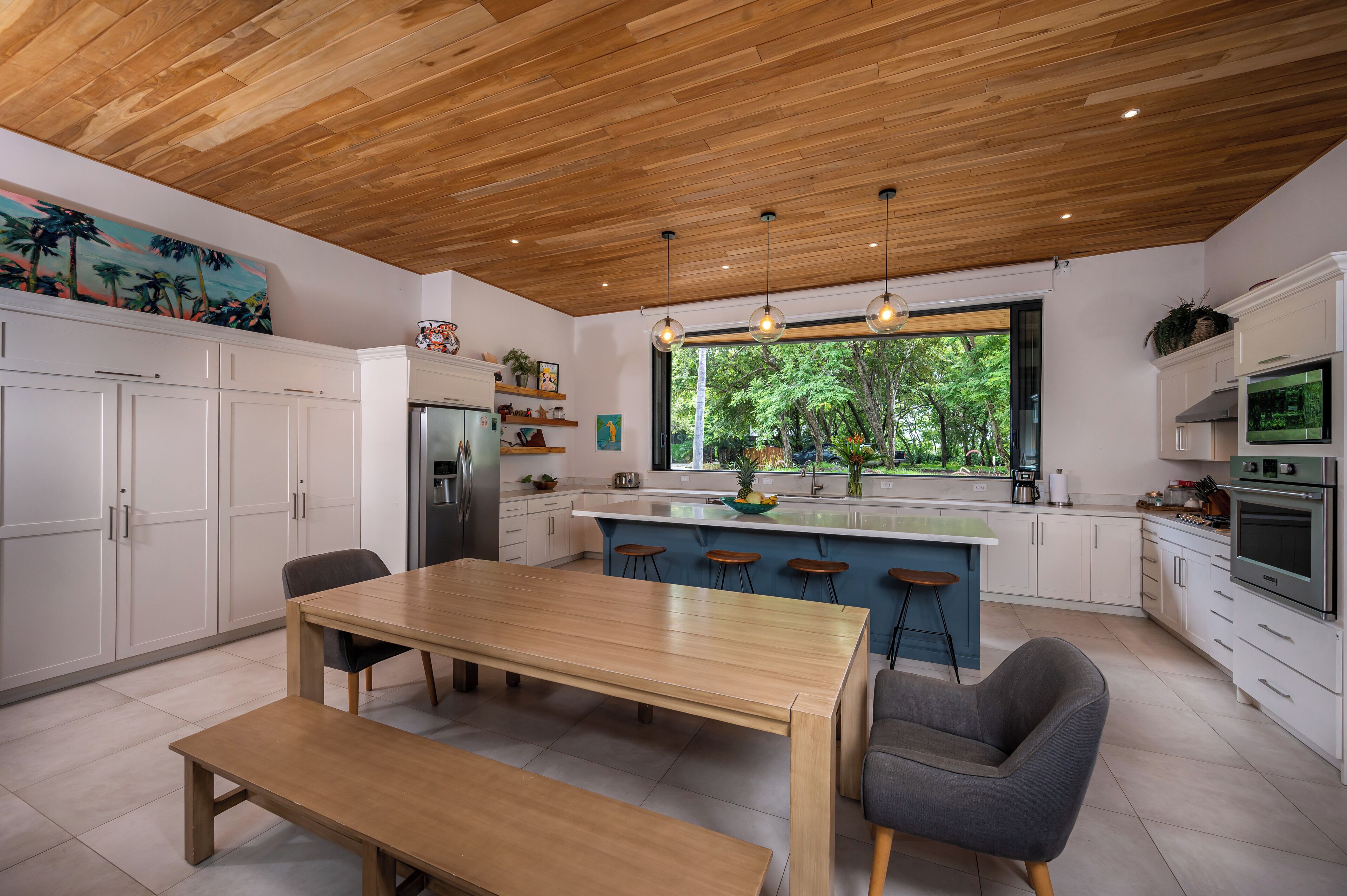 A modern kitchen with wooden ceiling, white cabinets, a central island with stools, and a large window offering a view of greenery.