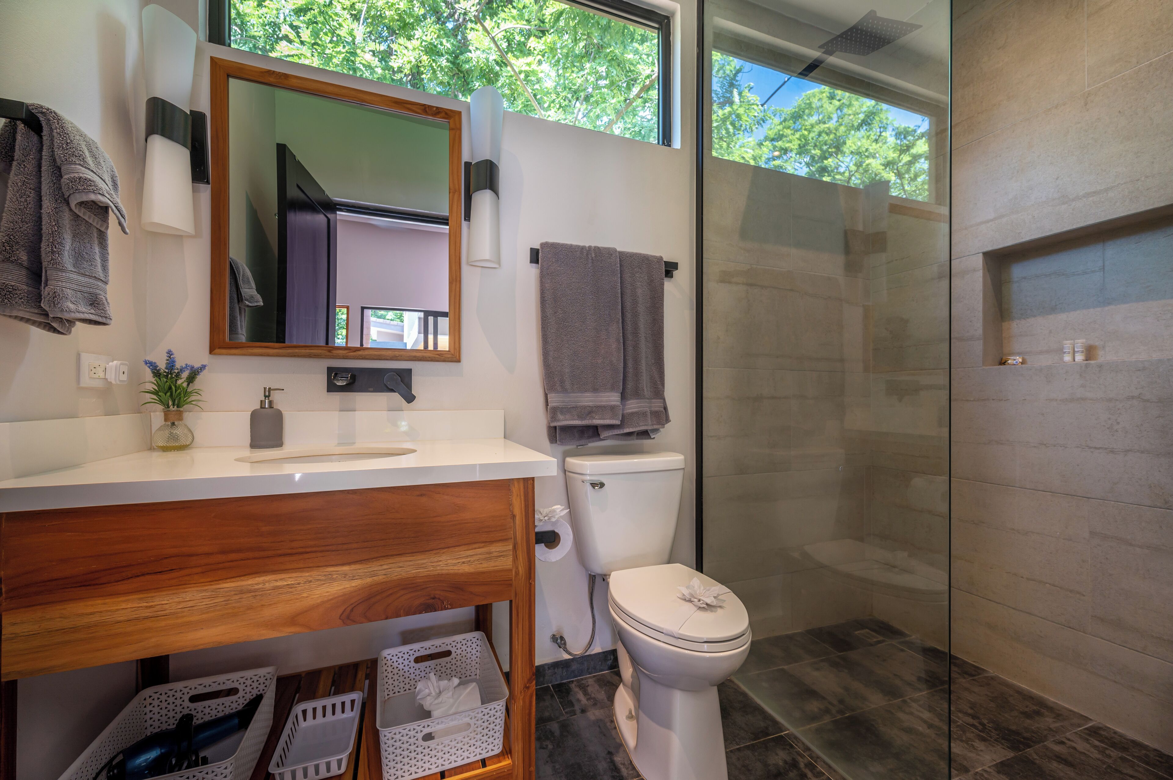 Un baño moderno con tocador de madera, inodoro, cabina de ducha con mampara de cristal y un ventanal que proporciona luz natural.
