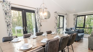 A dining area with a wooden table set for six, surrounded by grey upholstered chairs. There is a blue sofa in the background, and large windows provide a view of greenery outside. - Mayflower (Lifton)
