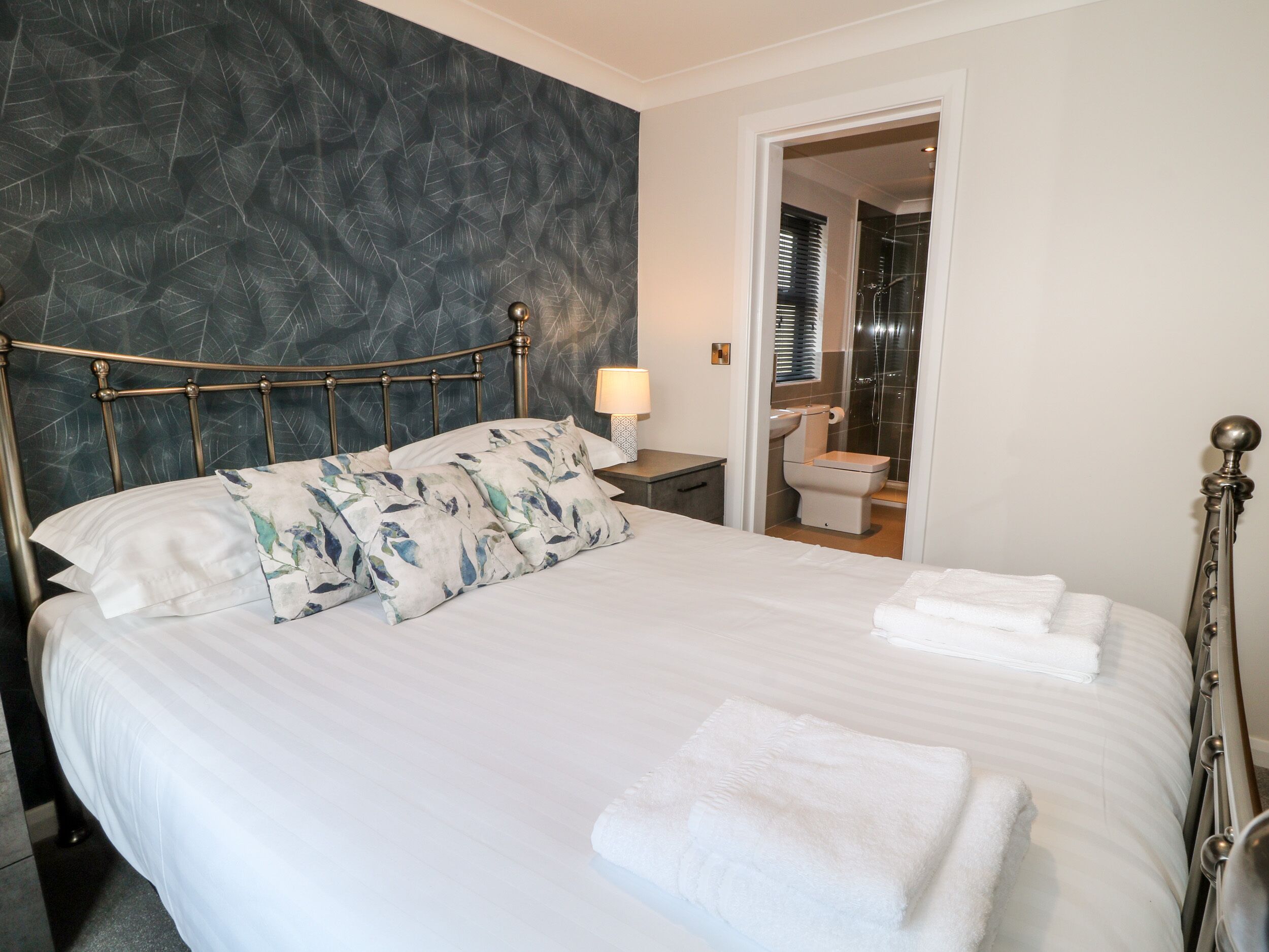A neatly made bed with white linens and floral pillows, a bedside table with a lamp, and a bathroom visible through an open door.