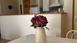A white vase with purple flowers on a table in a room with a kitchen in the background. - Gîte 2/4 people with swimming pool and view of the Orb valley (Cessenon sur Orb)