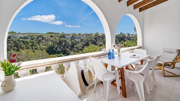 A balcony with a white table and chairs, a wicker chair, and a view of a forested landscape.