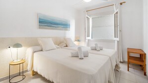 A hotel room with two beds, a ceiling fan, a painting, a bedside lamp, and a wooden side table.