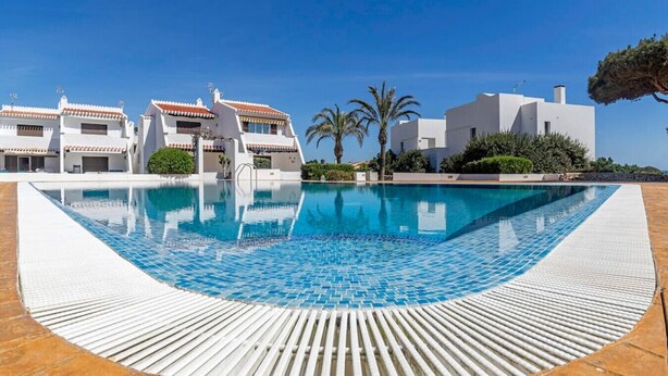 Una piscina con plataforma de baldosas, rodeada de edificios blancos con techos de tejas rojas, palmeras y un cielo azul despejado.
